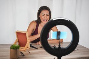 A young Hispanic female sits in front of a ring light while filming a TikTok video make-up tutorial